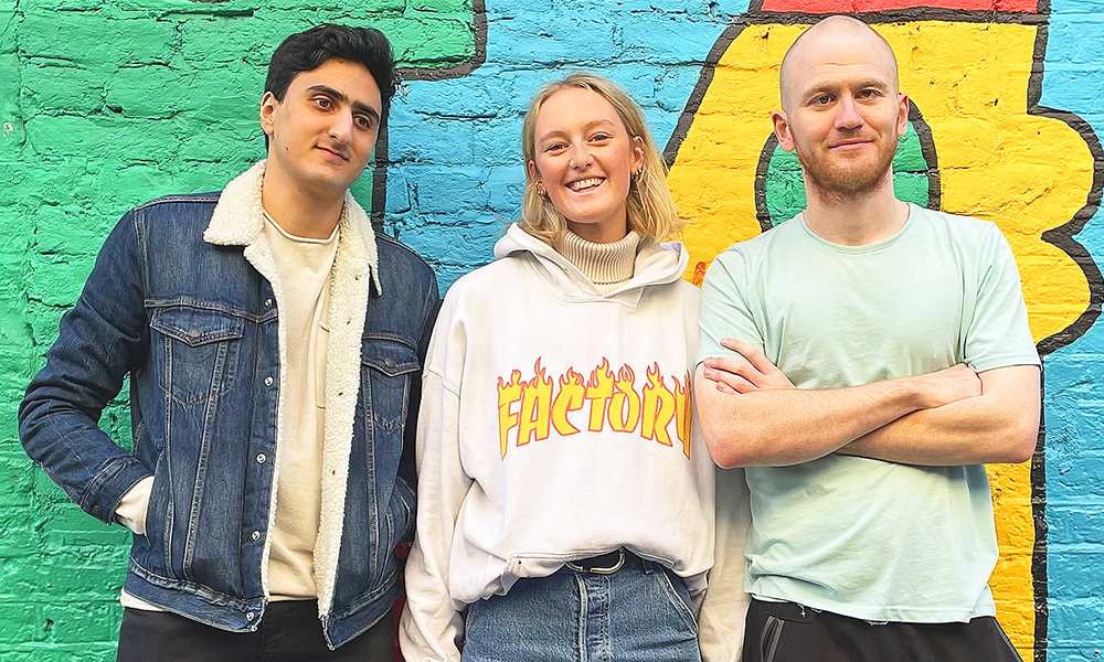 Image of FlawBored standing against a colourful wall, from left to right, Aarian Mehrabani,(6ft middle eastern man with thick black hair and a shaved face) Chloe Palmer (5ft 8” white woman with blonde hair, blue eyes and freckles.) and Sam Brewer (6ft 1” white man with a shaved head and a short ginger beard) 