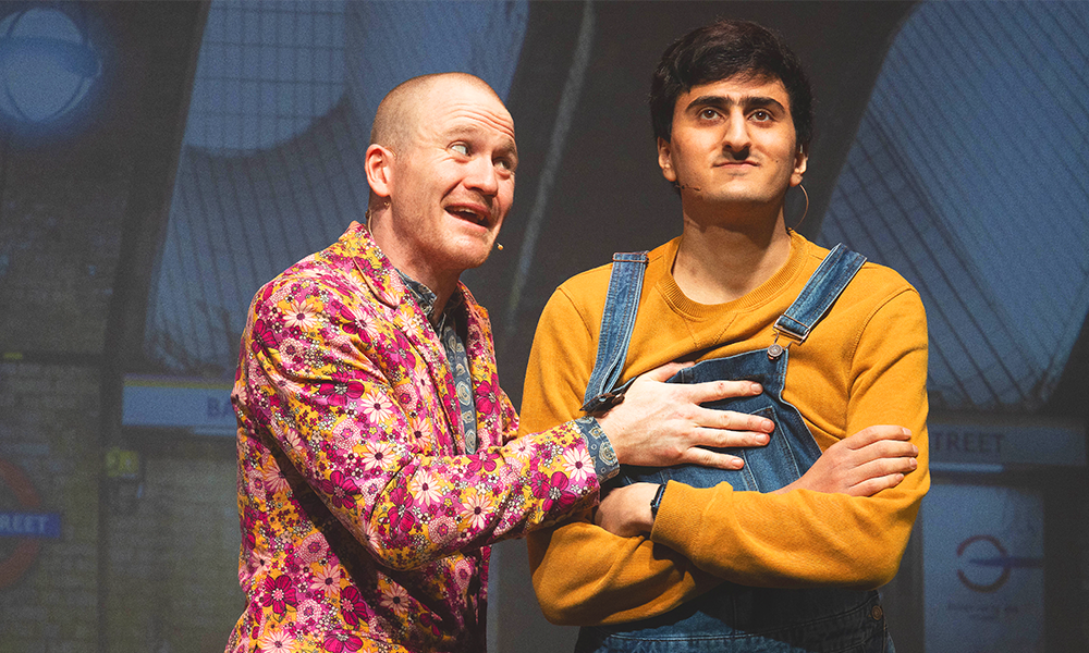 An image of Sam (left), wearing a pink floral suit jacket, holding Aarian (right), wearing a mustard jumper and blue dungarees both on stage. A photo of Baker Street tube platform projected behind