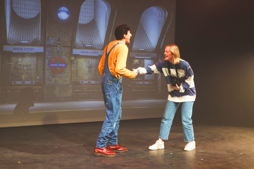 A photo of Aarian (left) and Chloe (right) on stage. Chloe (wearing a blue and white stripy jumper with blue jeans) extends her hand to Aarian (wearing blue denim dungarees and a mustard yellow jumper) A projection of Baker Street Tube Station in the background