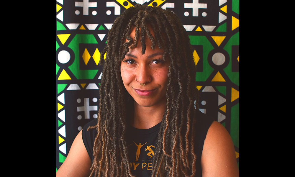 Chloe Redmond, entrepreneur and owner of The Body People – a young woman with long dreadlocks in a black top in front of a green, gold, black and white geometric wax print fabric