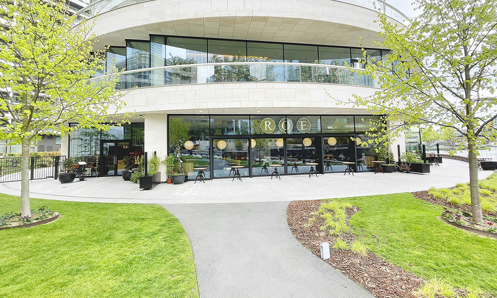 Image shows the exterior of Roe restaurant at the base of the One Park Drive tower in Wood Wharf. It has a large terrace and overlooks the waters of nearby South Dock