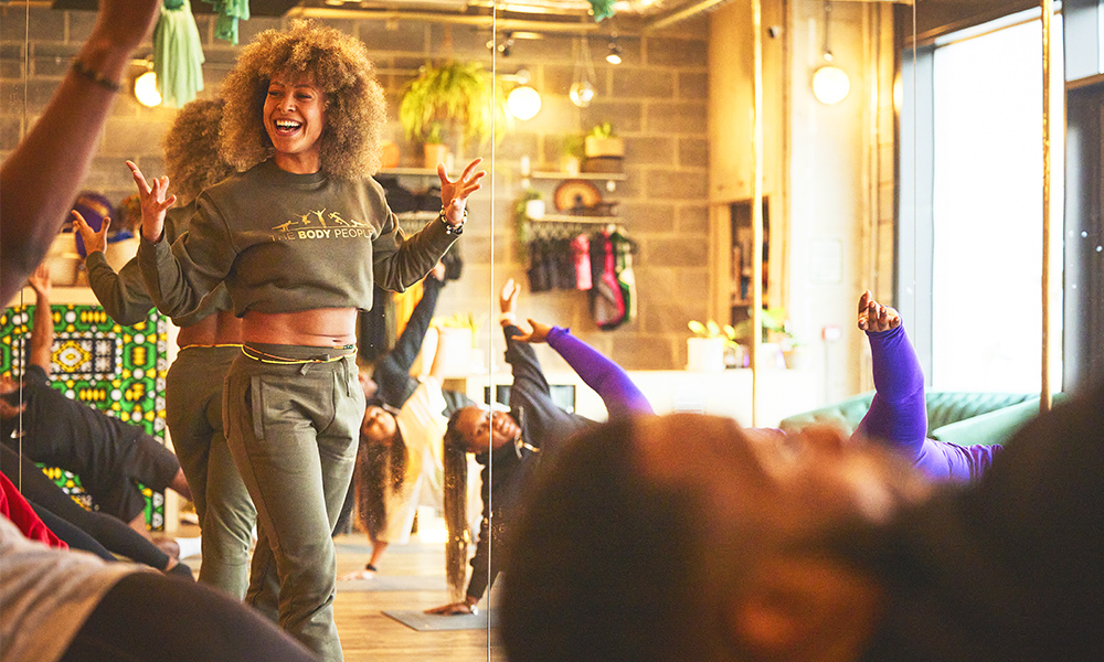 Chloe Redmond, wearing a khaki tracksuit with a crop top raises her hands in a fitness studio while participants practise Pilates movements on the mat