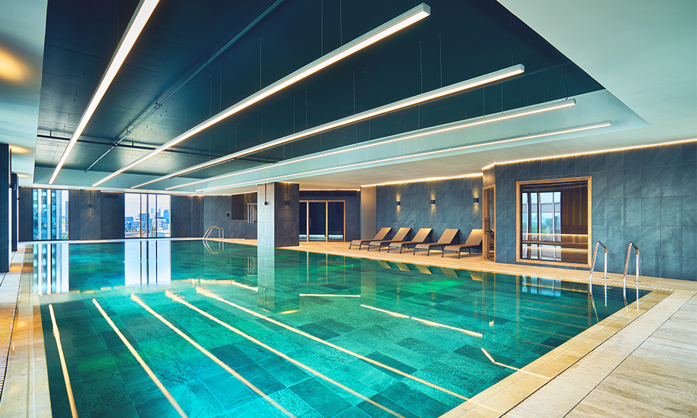 Image shows a bright turquoise swimming pool surrounded by light brown limestone times at Third Space Wood Wharf
