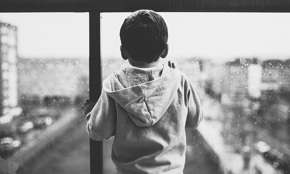An image of a lonely boy looking out from a rain-streaked glass balcony