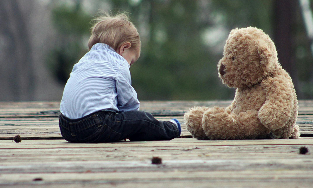 A boy sits alone with a teddy bear on some wooden planks