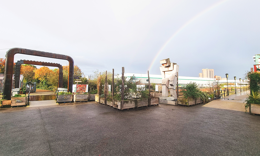 Image shows a general view of Cody Dock on the River Lea under a rainbow with a toothed rolling bridge made of iron and a large wooden sculpture of a figure