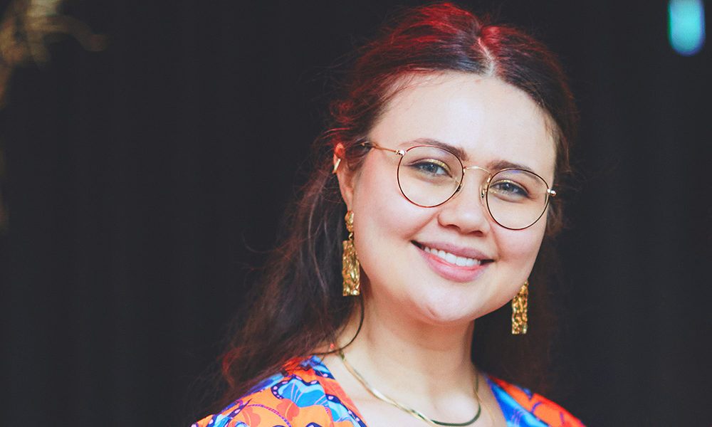 Image shows artist, illustrator and Power Of Women founder Leah Sams, a dark haired woman in glasses with gold earrings wearing one of her orange dresses