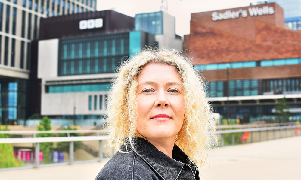 Image shows Tamsin Ace, a woman with curly blonde hair in a black denim jacket in front of buildings at Stratford's East Bank