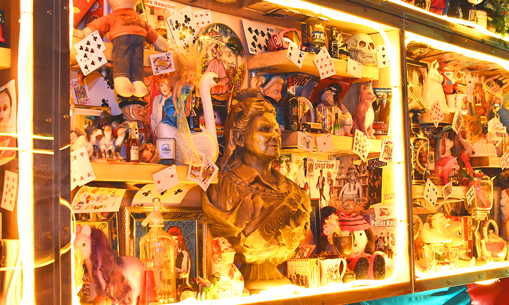 Image shows a lit cabinet filled with playing cards, toys, records and a bust of Pat Butcher in the style of Queen Victoria