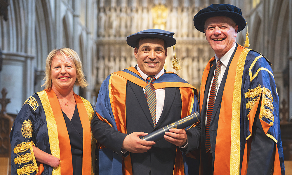 Images shows a man, Jonathan Ganesh, flanked by a woman and another man all in academic gowns, smiling at the camera