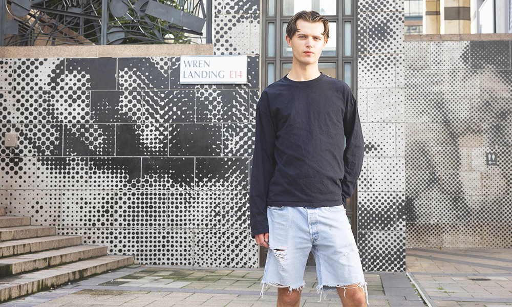 Artist Henry Gibbs stands in front of his mural Get Real in Canary Wharf, a young man in a black top and denim shorts