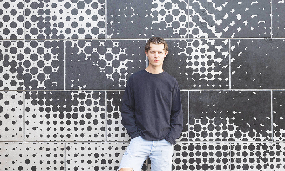 Artist Henry Gibbs stands in front of his mural Get Real in Canary Wharf, a young man in a black top and denim shorts