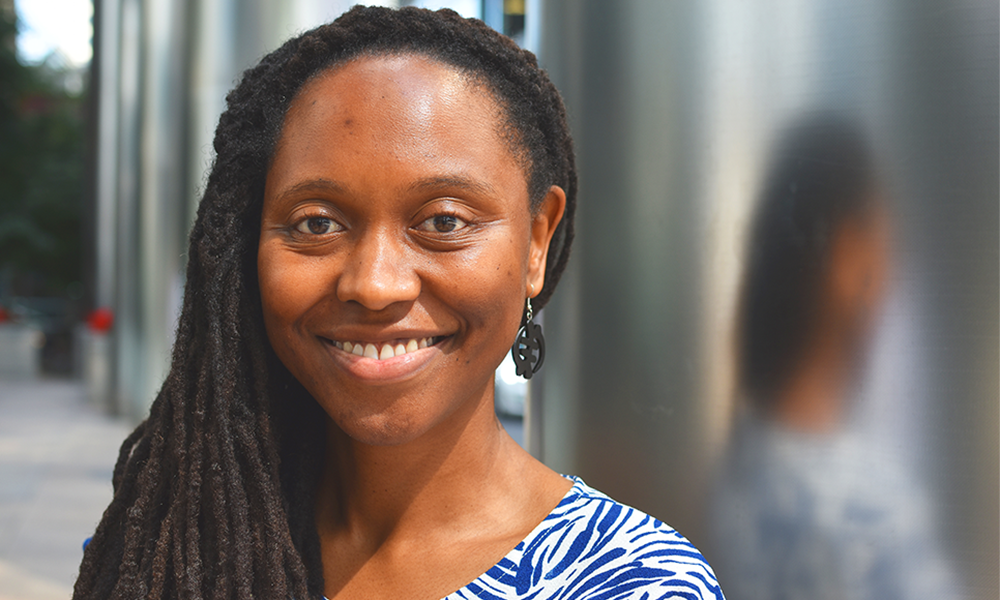 Image shows UCL School Of Management Behavioural Research Lab manager Sharmay Mitchell outside One Canada Square in Canary Wharf. A woman wearing a blue and white dress with long dreadlocks swept to the left