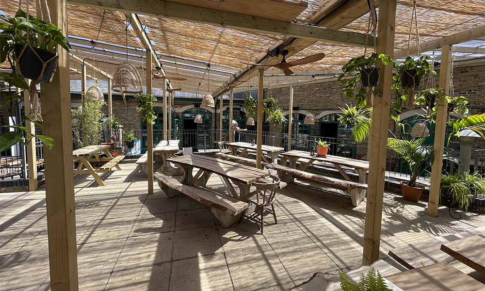 Image shows wooden tables under a wooden and plastic weatherproof pergola at the back of the pub