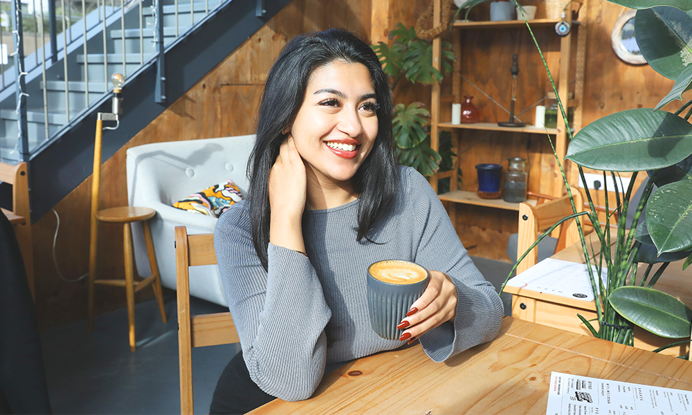 Image shows Farhana sitting in a wood-lined cafe enjoying a cup of coffee from The Well Bean Co