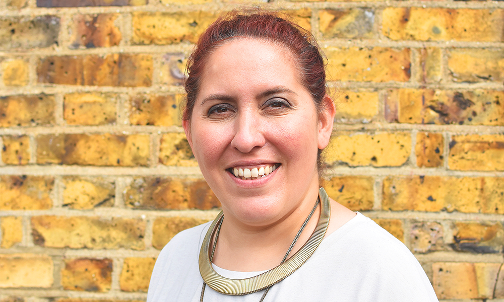 Image shows 3equals1 Design founder and director Maz Mahmoudi, a woman wearing a gold necklace with red hair in front of a yellow brick wall