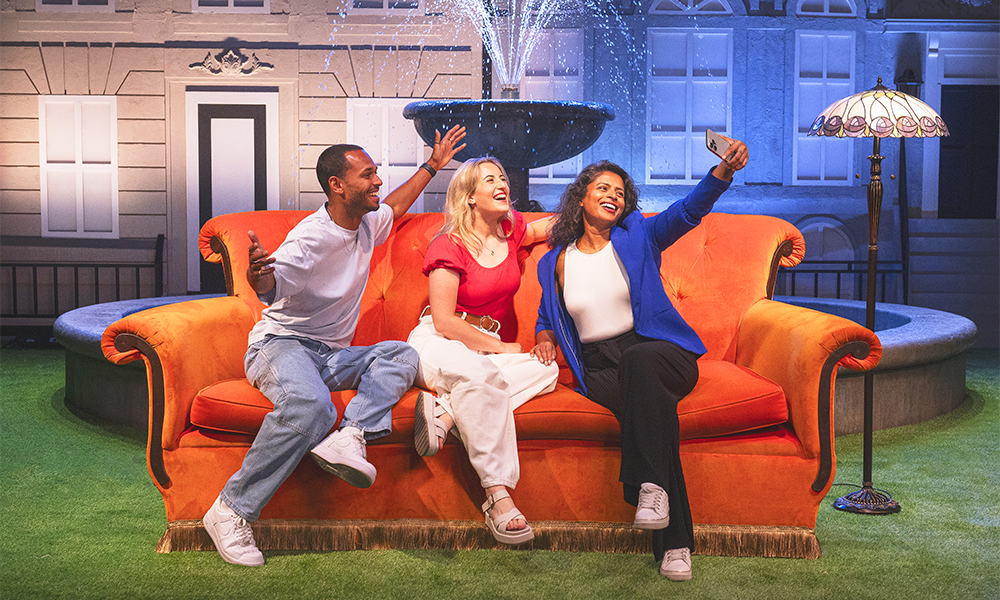 Three people sit on the orange Friends sofa in front of a fountain at Friends The Experience: The One In London