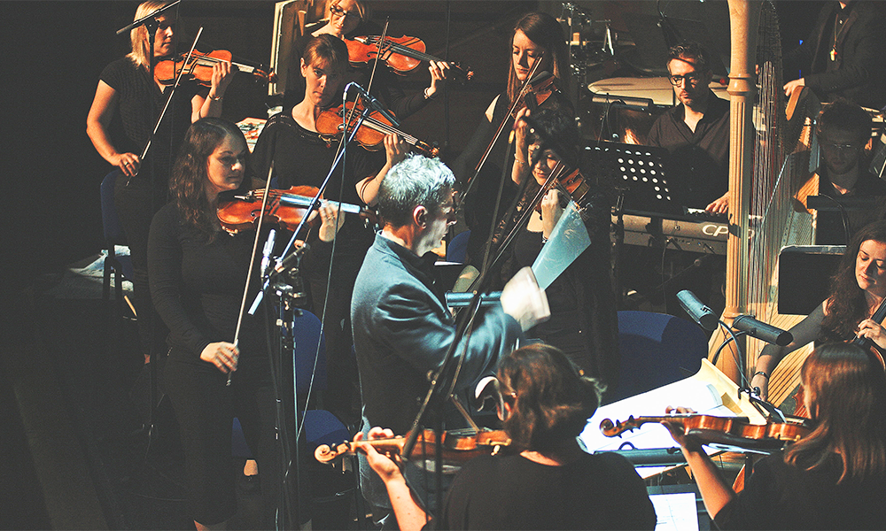 Image shows Spencer Down conducting the string section of the Docklands Sinfonia with a keyboard player and harpist in the background