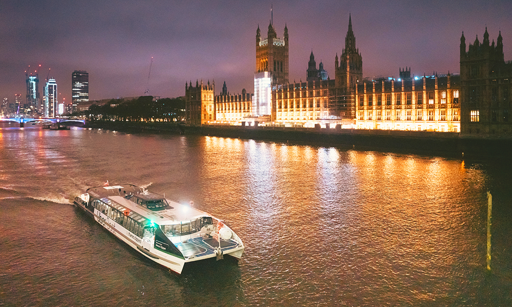 Image shows an Uber Boat By Thames Clippers river bus sailing past the houses of Parliament at Night