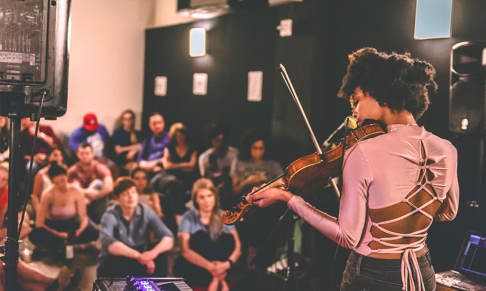 Image shows a Sofar Sounds performer, a violinist, playing to a crowd. The company has partnered with Uber Boat By Thames Clippers