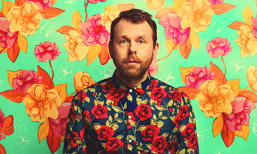 Image shows comedian, a man with short dark hair, in a bright floral shirt covered with roses