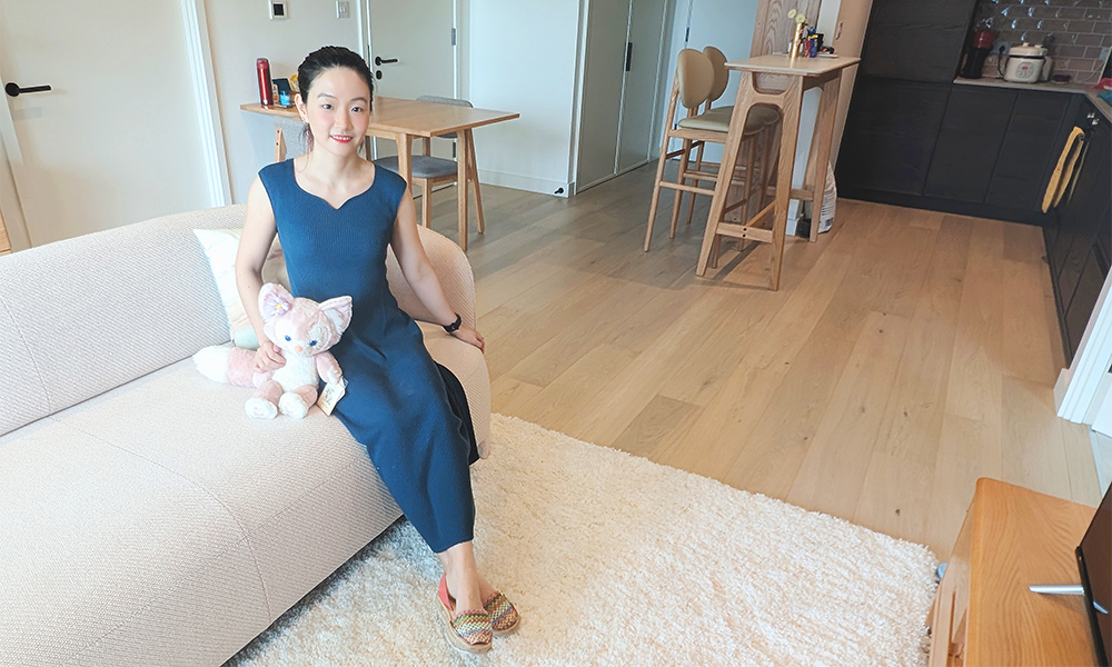 Image shows Riverscape buyer Sophie Gong in her east London apartment, a woman in a blue dress on a beige sofa with a stuffed cat