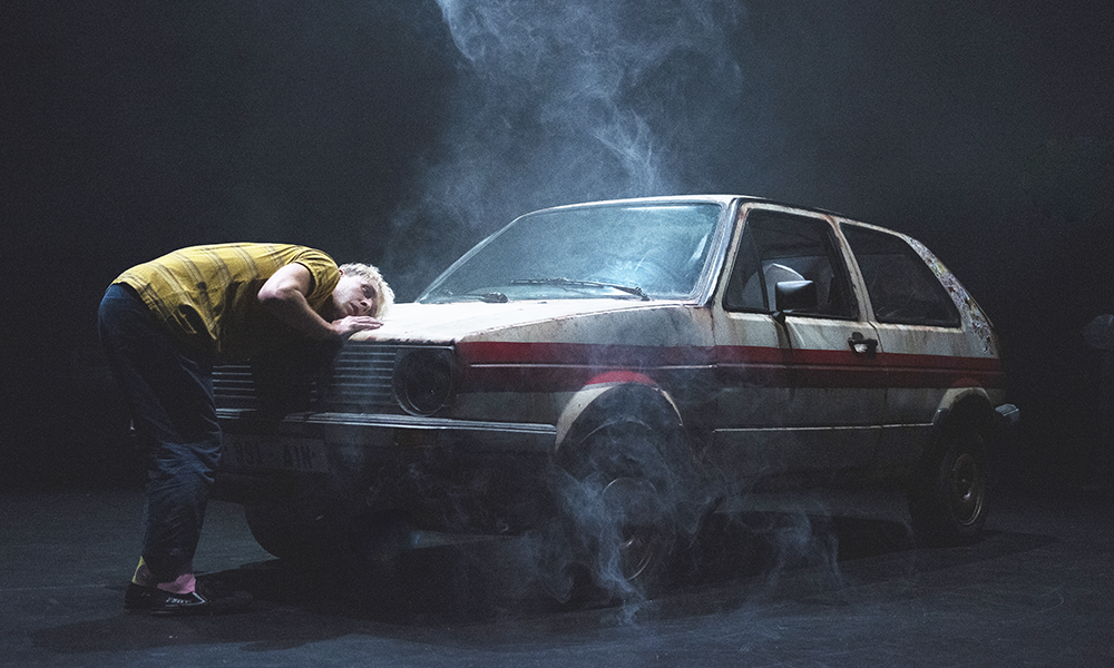 Image shows a dancer in Birdboy, a man bending over a smoking car